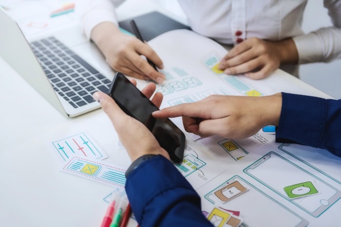 Zwei Personen bei einer Business-Beratung. Eine davon sitzt am Laptop, die andere zeigt auf ein Handy.