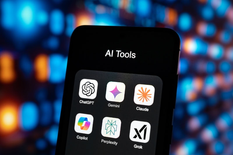 Smartphone mit App-Logos von KI-Tools wie ChatGPT, Gemini, Claude, Copilot, Perplexity und Grok auf dem Display.