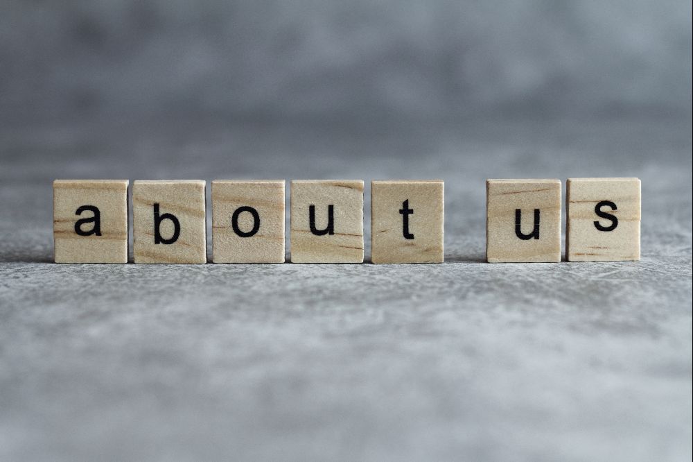 Holzwürfel mit schwarzen Buchstaben bilden den Schriftzug „about us“ auf grauem Hintergrund.