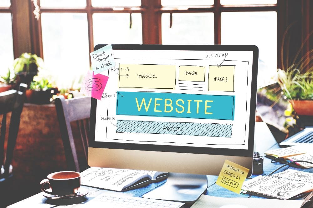 Computerbildschirm mit einer Skizze für den Aufbau einer Website, umgeben von Notizen, Büchern und einer Kaffeetasse auf einem Schreibtisch.